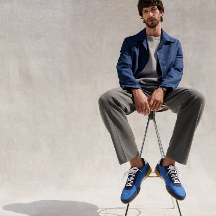 Man sitting on a stool wearing a blue jacket, gray pants, and blue sneakers.