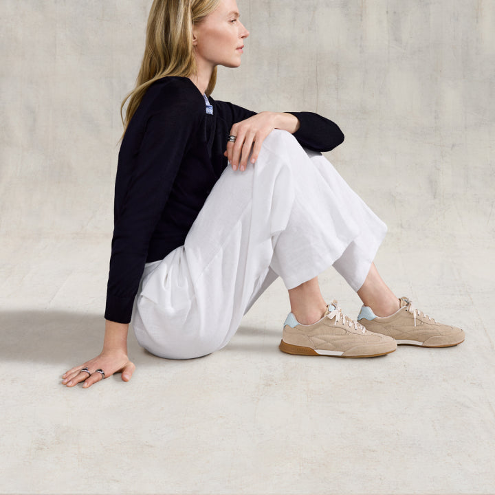Woman sitting on a light-colored floor wearing a black top, white pants, and beige sneakers.