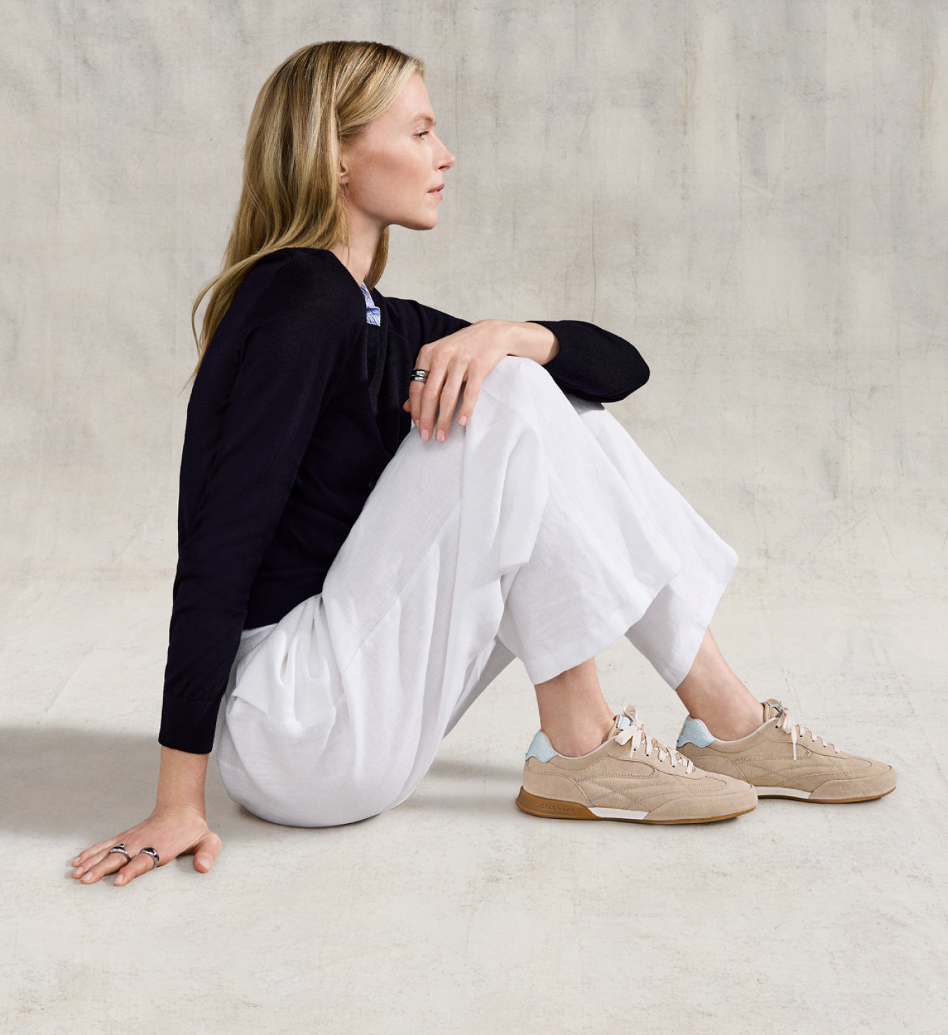 Woman sitting on the floor wearing a black top, white pants, and beige sneakers.