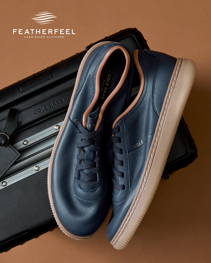 A pair of navy blue leather low-top sneakers with beige soles and light tan lining, displayed on top of a black suitcase against a warm brown background. The Featherfeel brand logo is visible in the top left corner.