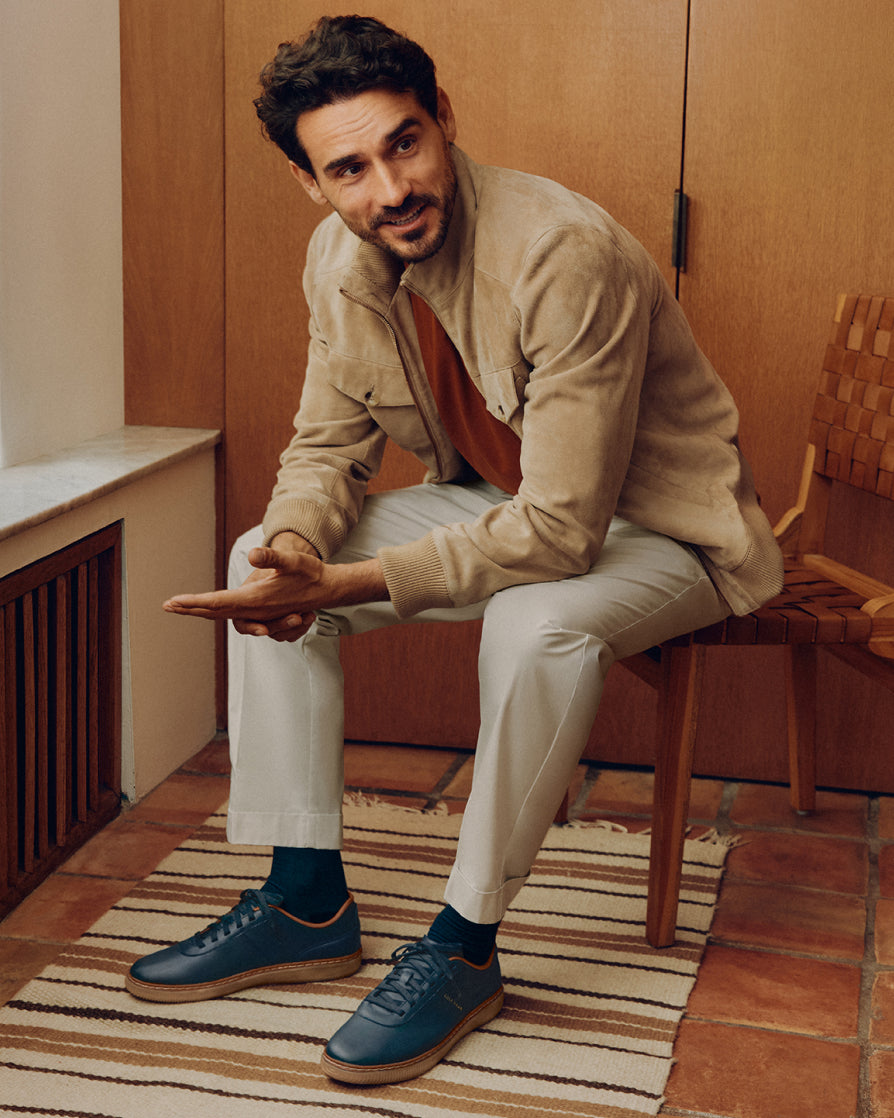 A man sits on a wooden chair in a warm indoor setting, wearing a beige jacket, light trousers, dark blue shoes, and dark socks.