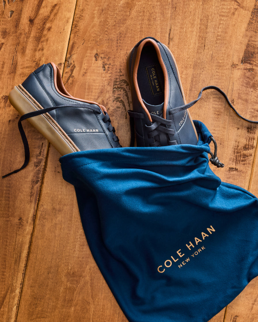 A pair of dark blue leather Cole Haan lace-up shoes with tan soles, arranged on a wooden floor next to a blue drawstring dust bag with gold lettering.