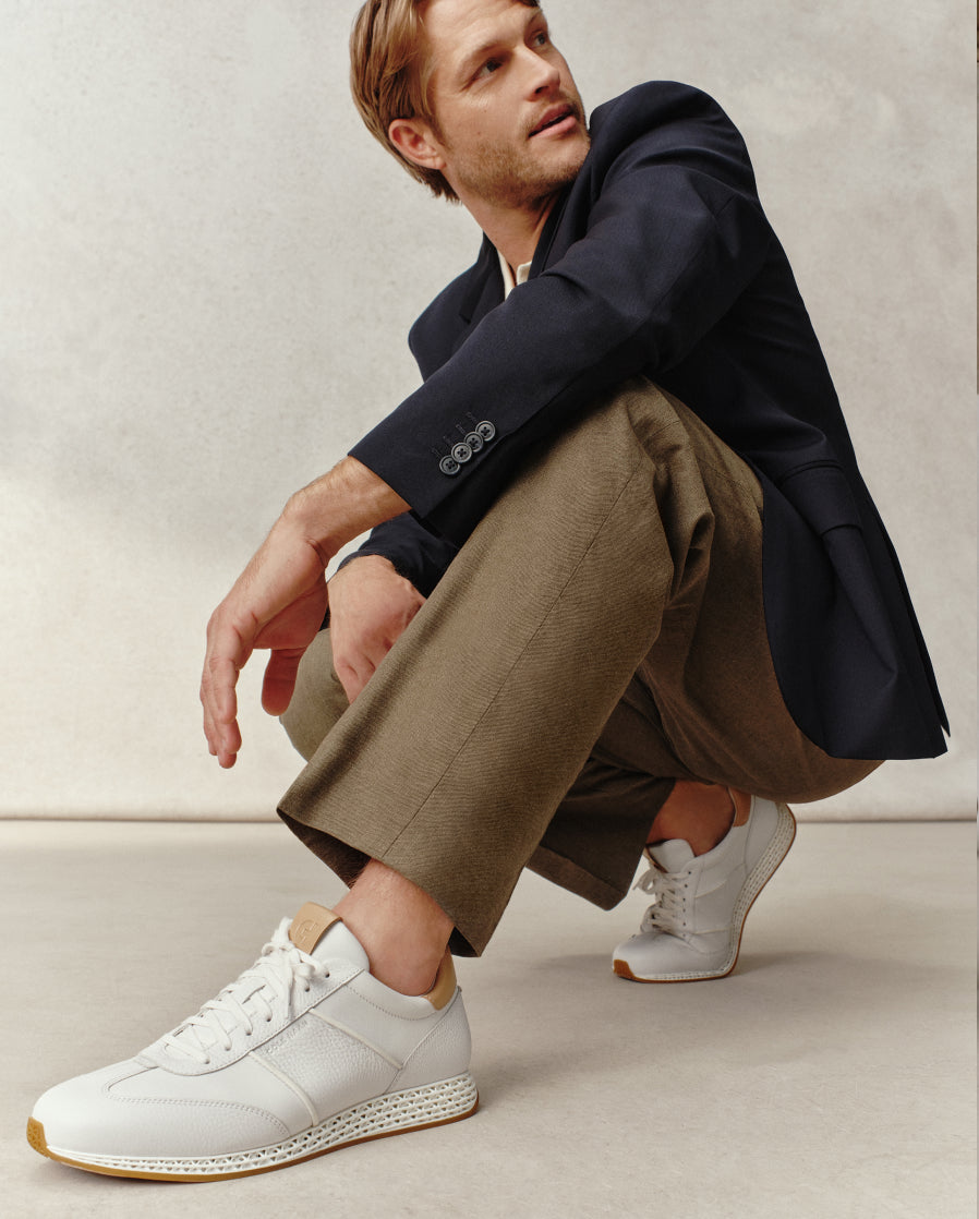 Man wearing a navy blazer, beige pants, and white sneakers on a neutral background