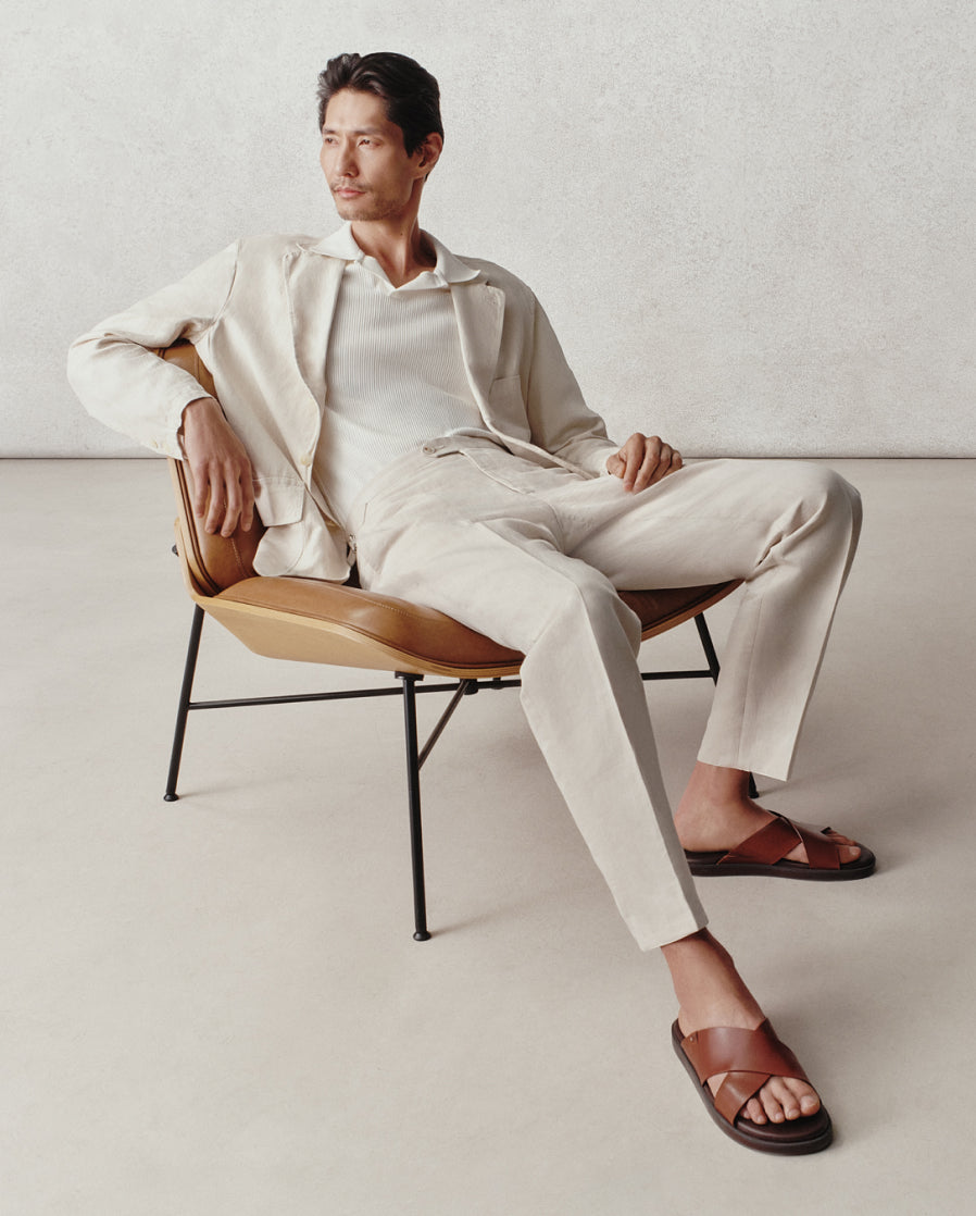 Man sitting on a chair wearing a white outfit with brown sandals in a minimalistic setting.