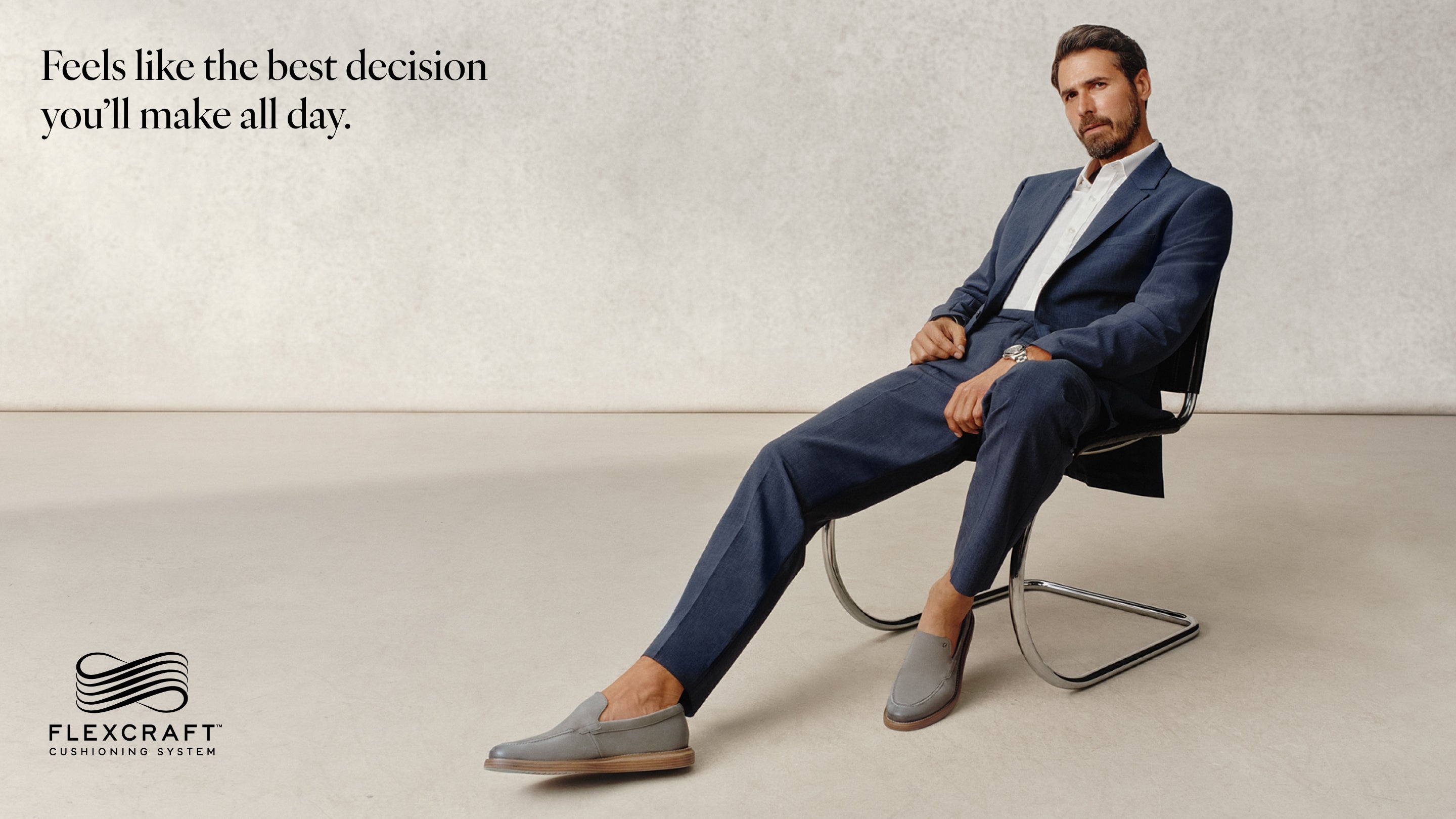 Man in a blue suit sitting on a chair with a neutral background, wearing loafers