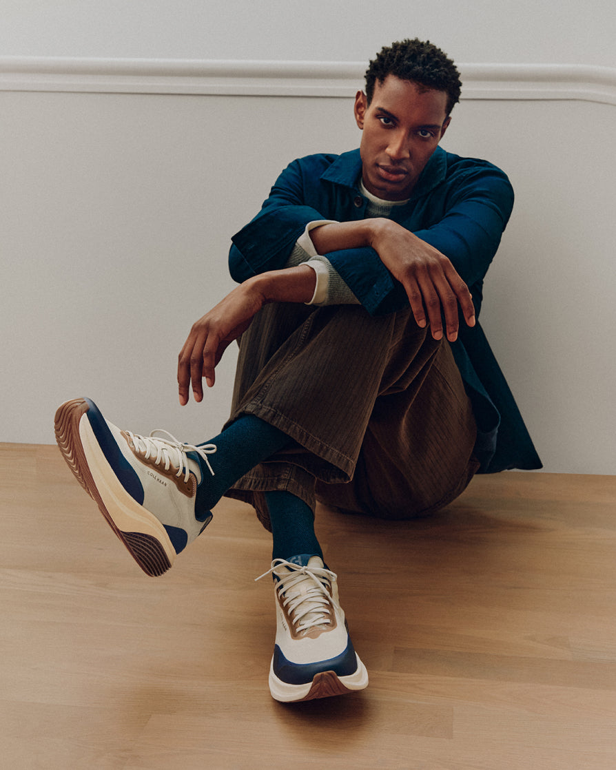 A man sits on a light wood floor against a white wall, wearing a dark blue overshirt, brown corduroy trousers, dark blue socks, and cream and navy running sneakers.