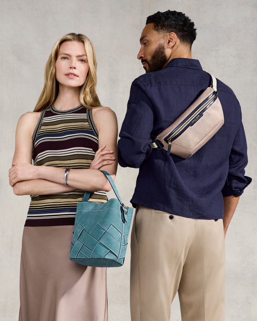 Man and woman standing close together, with the man wearing a beige crossbody bag and the woman holding a blue woven handbag.