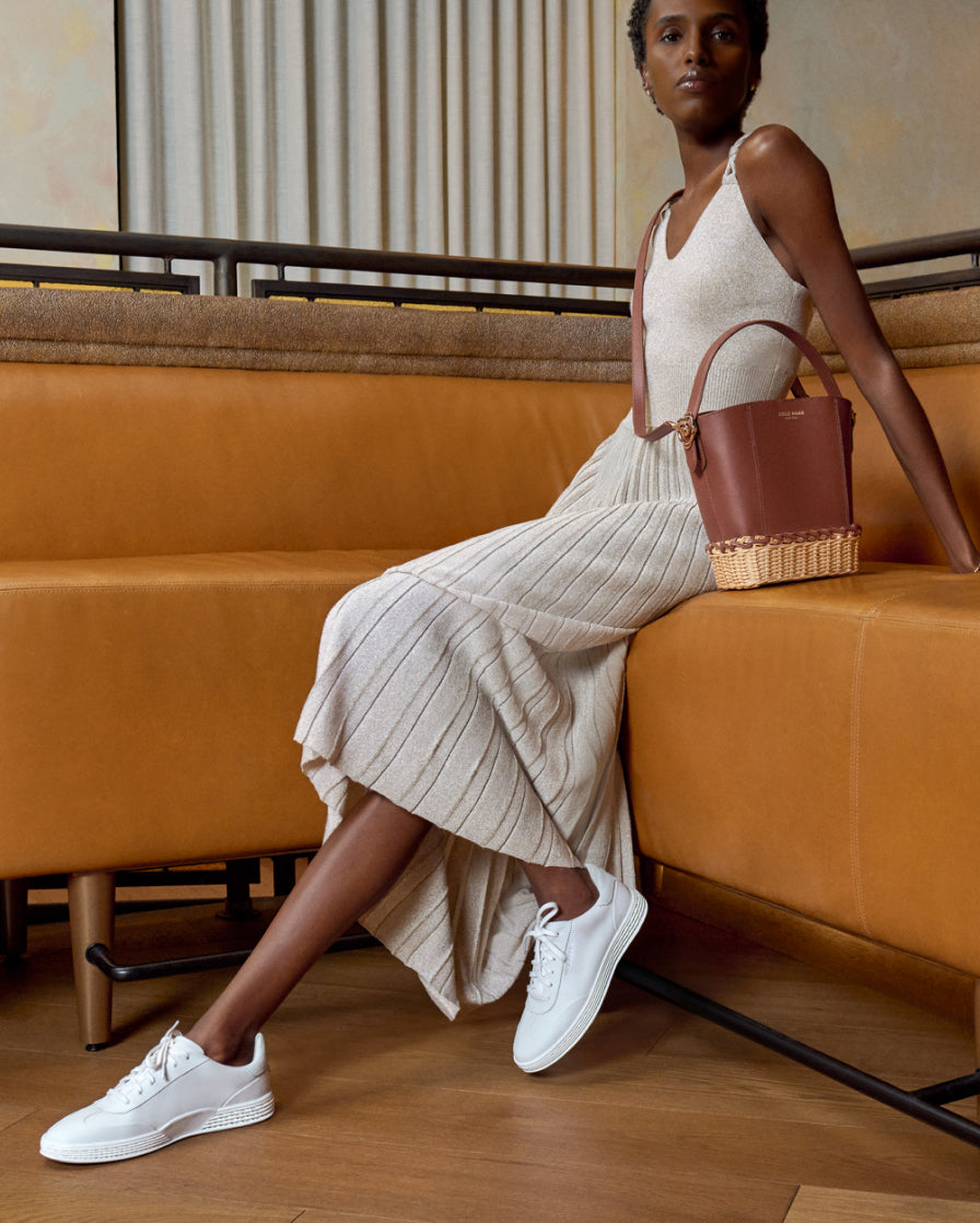 Woman sitting on a mustard yellow couch wearing a beige dress, white sneakers, and holding a brown handbag.