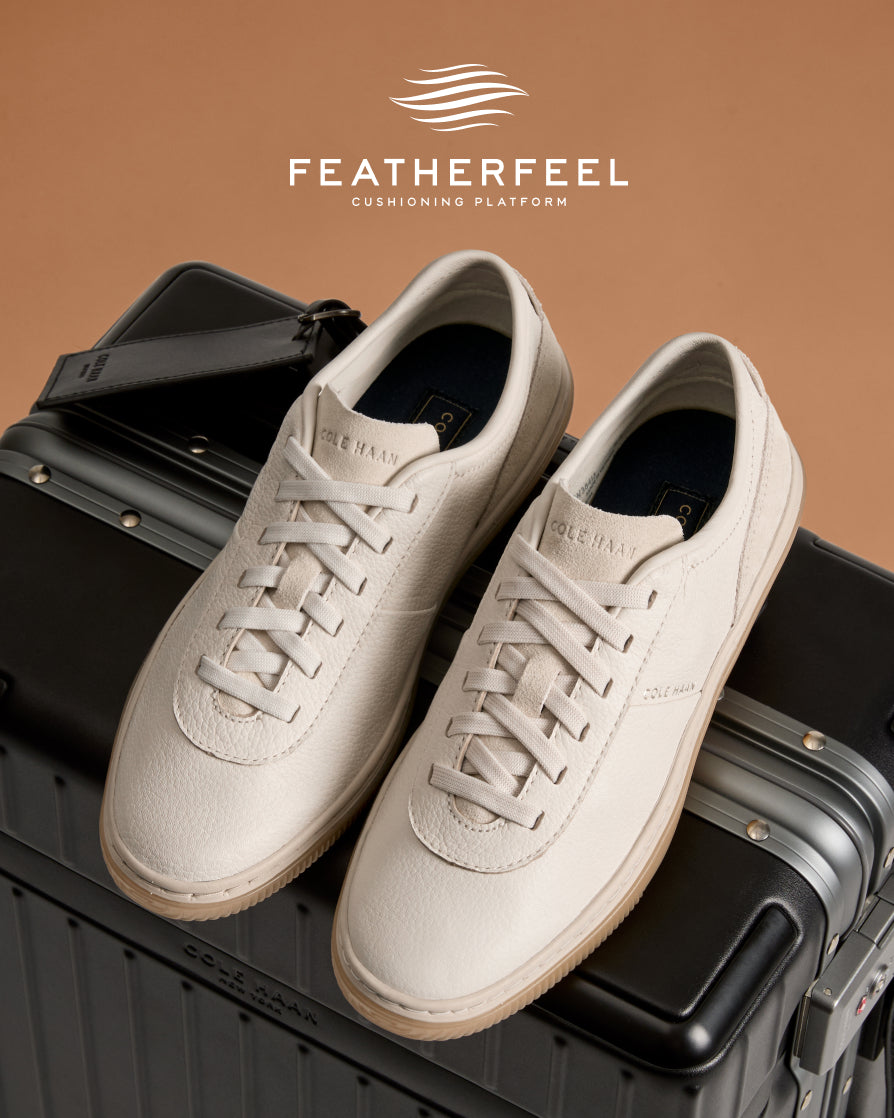 A pair of off-white leather low-top sneakers, styled on top of a black hard-shell suitcase against a warm tan background.