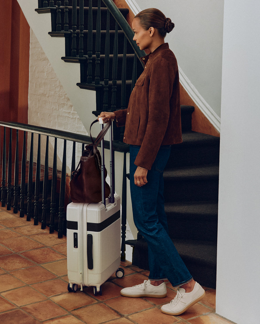 A woman in a brown suede jacket, blue jeans, and white sneakers stands next to a staircase holding a white rolling suitcase with a brown leather bag on top.