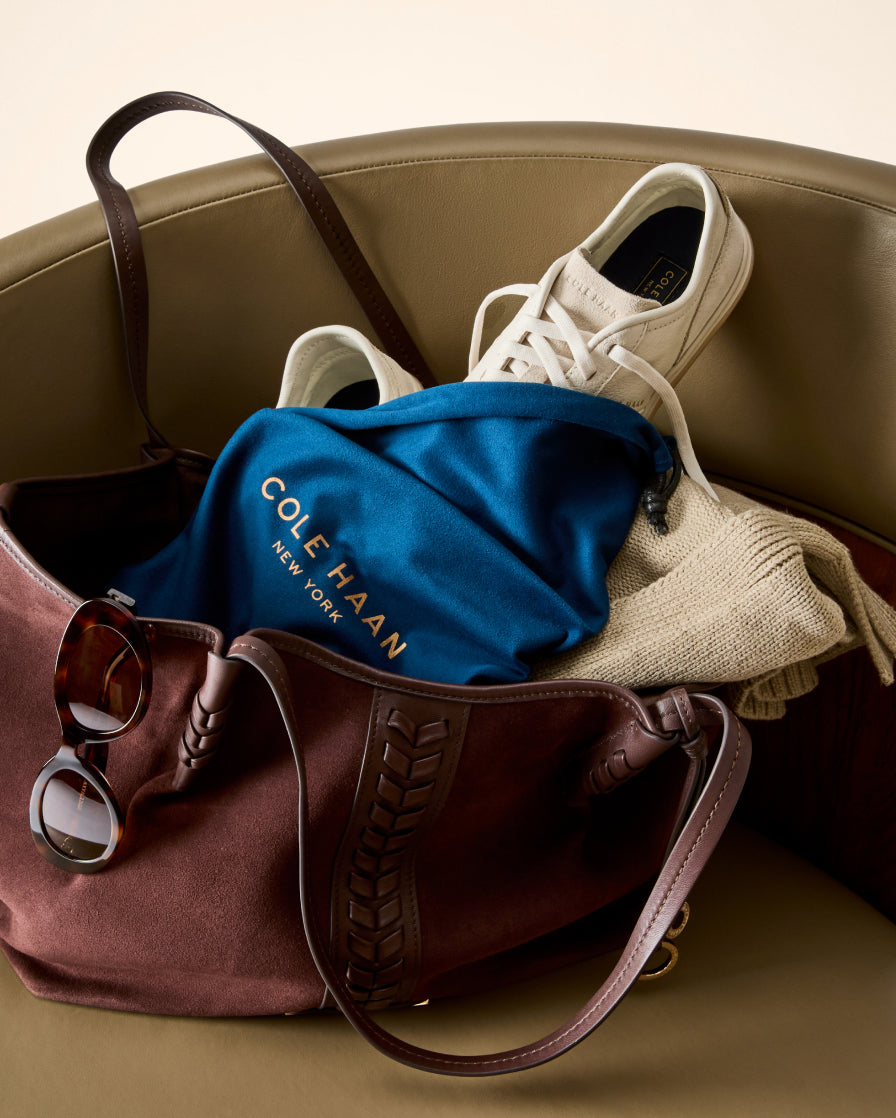 A brown leather tote bag filled with a blue Cole Haan New York bag, a cream knit sweater, white sneakers, and sunglasses, set against a beige leather chair.