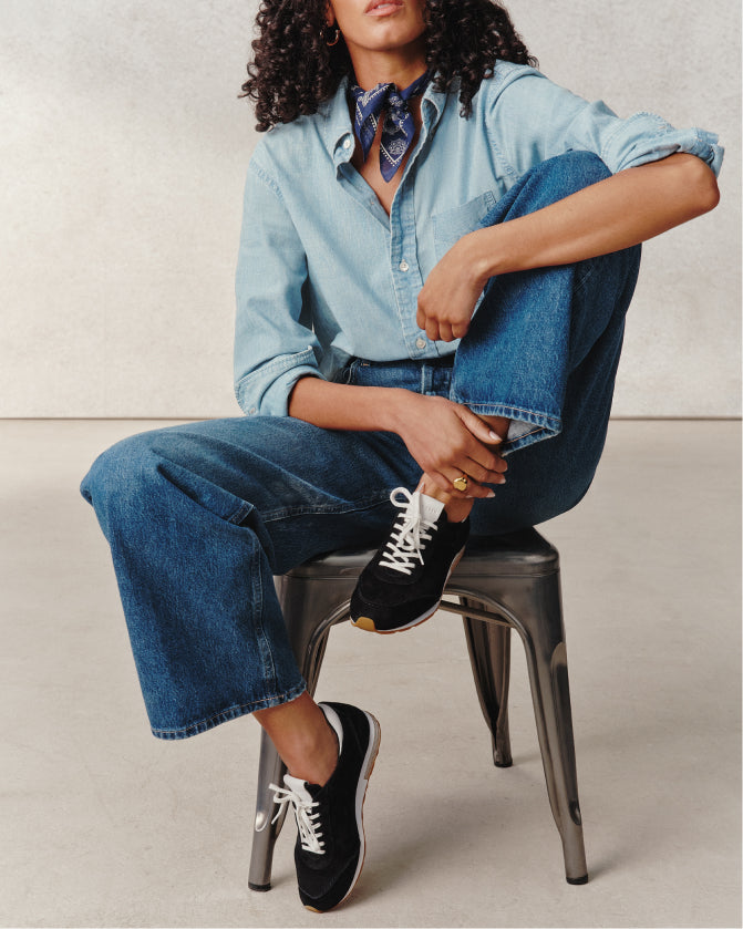 Person wearing a denim outfit with black sneakers sitting on a stool.