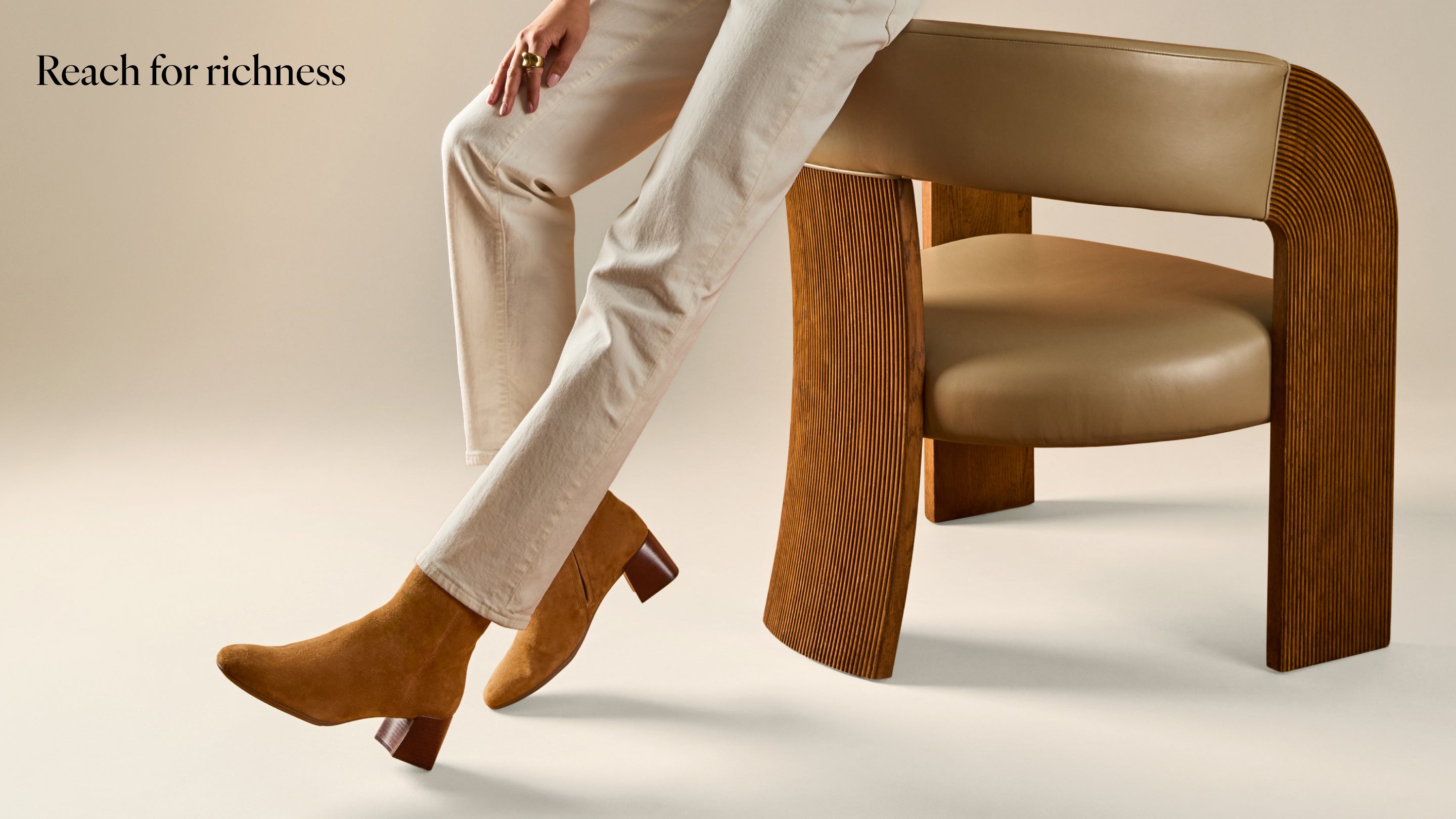 A person seated on a modern wooden armchair wearing light beige trousers and tan ankle boots.