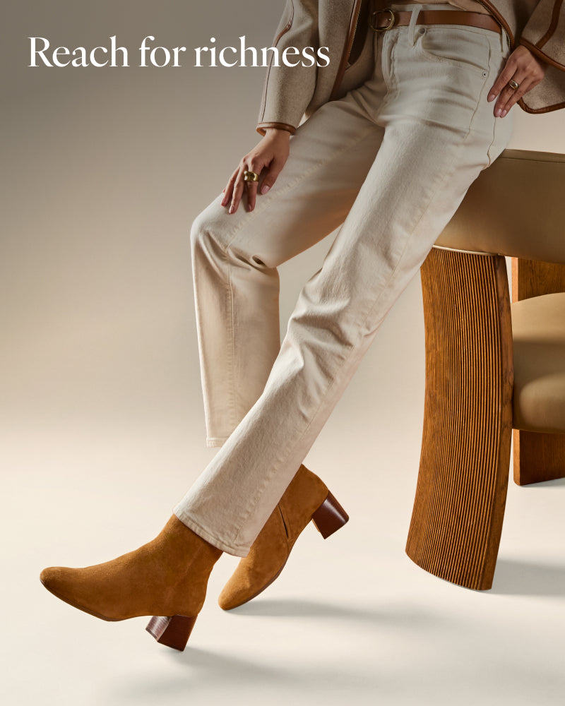 A model seated on a wooden chair wearing light beige straight-leg jeans and tan suede ankle boots.