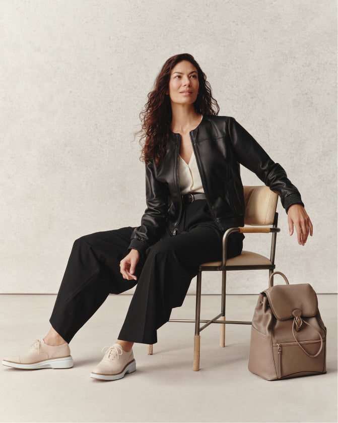Woman in black leather jacket and pants sitting on a chair with a beige bag beside her.