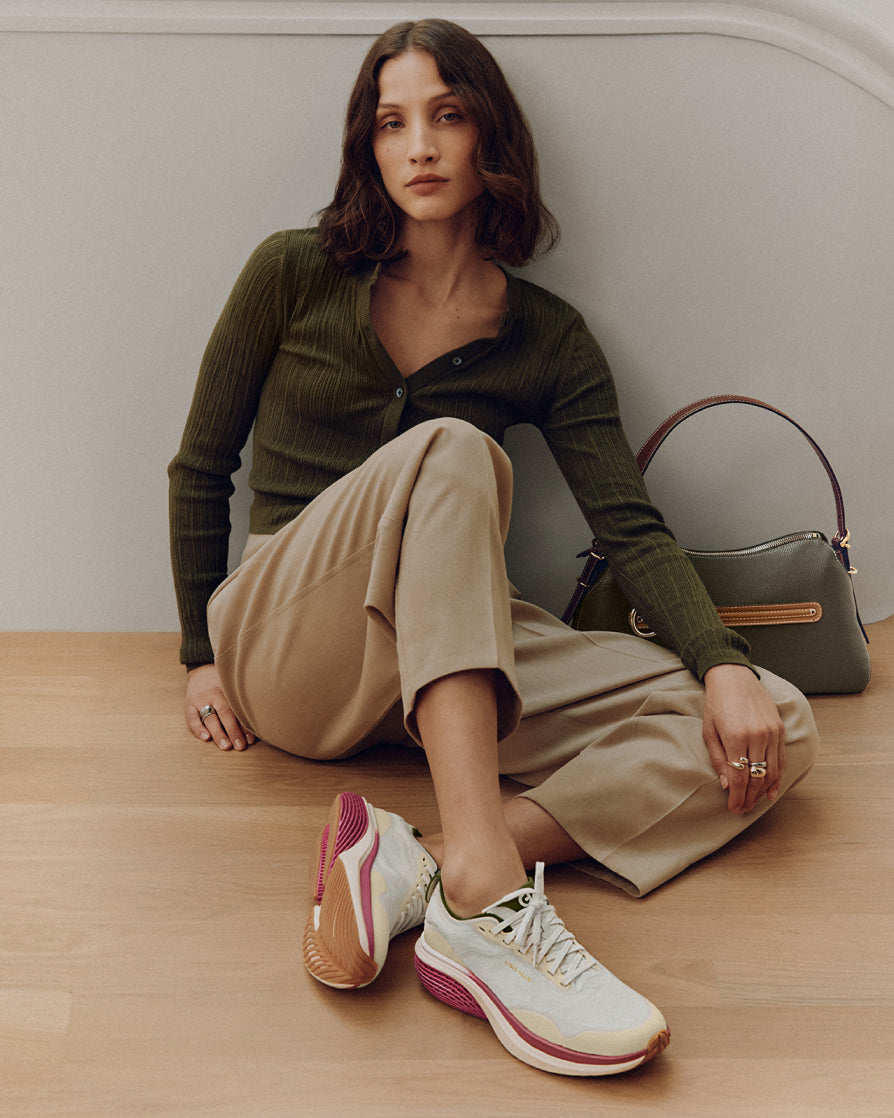 A woman sits on a light wood floor against a neutral gray wall, wearing an olive green ribbed cardigan, beige wide-leg cropped trousers, and white sneakers with pink and gold accents. A matching olive green handbag rests beside her.