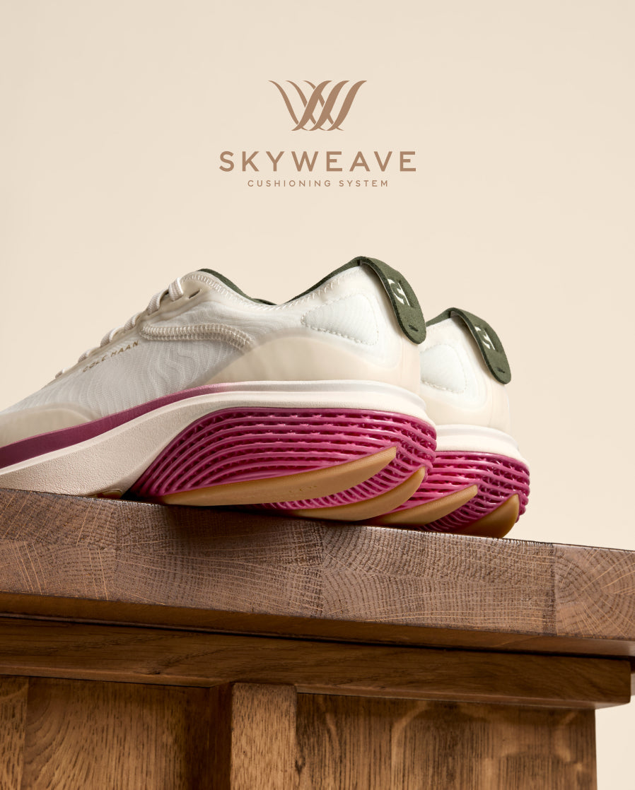 A pair of white sneakers with magenta-pink midsoles and dark green heel accents, resting on a rustic wooden block against a beige background.