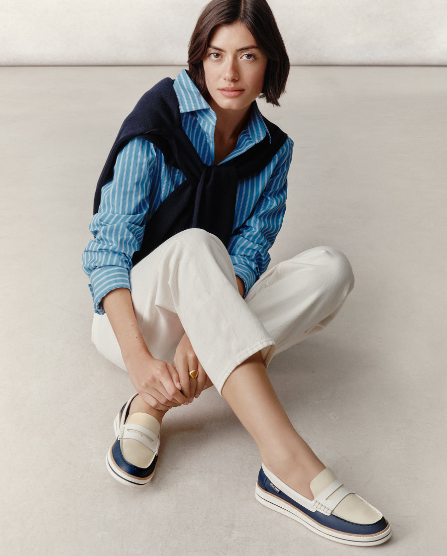 Woman wearing a blue striped shirt, white pants, and navy loafers sitting on a light-colored floor.