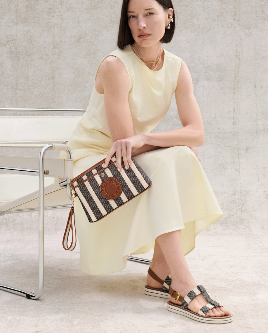 Woman in a light yellow dress holding a striped clutch bag, sitting on a chair.