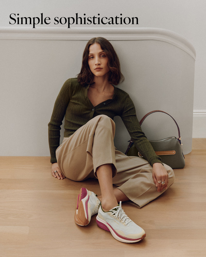 A woman sits on a light wood floor against a white curved wall, wearing an olive green ribbed long-sleeve top, beige wide-leg trousers, and white sneakers with pink and tan accents. A shoulder bag sits beside her