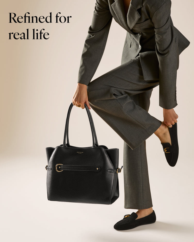 A model in a gray suit holding a black leather handbag with gold hardware, wearing black loafers