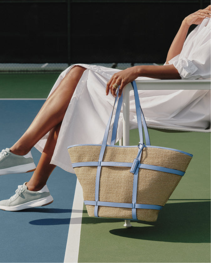 Person sitting on a tennis court holding a straw bag with blue straps